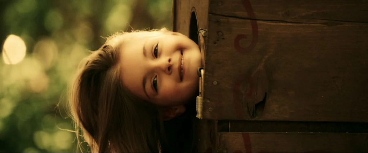 Kiss Kiss Bang Bang (2005) movie still: closeup — A wooden box structure sits outdoors with blurry green foliage in the background. A young girl…