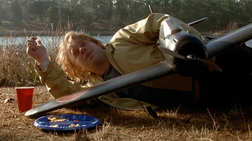 Love Liza (2002) movie still: medium — A man lies on dead grass beside a lake in a rural area. A red plastic cup and a blue plate sit on the ground…
