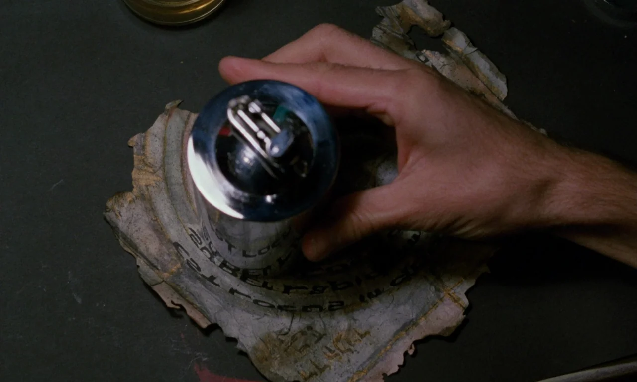 The Church (1989) movie still: closeup — A metallic cylindrical object rests on a piece of torn, weathered, printed parchment. A hand is positioned…