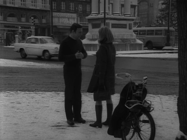 My Night At Maud’s (Ma Nuit Chez Maud) (1969) movie still: fullbody — A snowy public square features a stone monument and a passing vintage…
