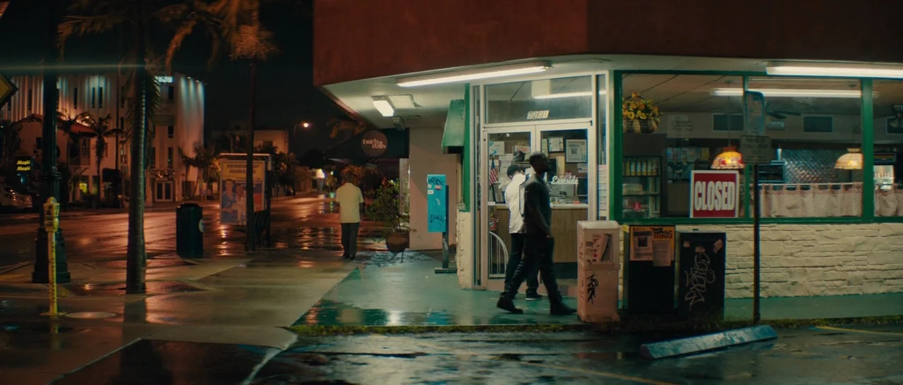 Moonlight (2016) movie still: wide — A nighttime urban scene with a wet, reflective sidewalk outside a closed retail establishment. Two men are…