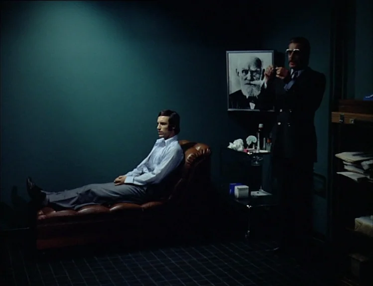The Arrival of Joachim Stiller (1976) movie still: fullbody — A dimly lit room features a leather chaise longue against dark blue-green walls. A man…