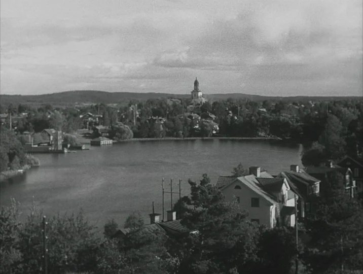 Crisis (1946) movie still: establishing — A quiet lakeside town features several white houses nestled among dense trees and vegetation. A prominent…