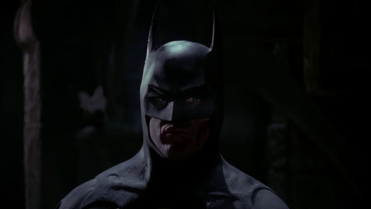 Batman (1989) movie still: closeup — A dark, dimly lit environment provides the backdrop for a close portrait. The masked figure stares forward with…