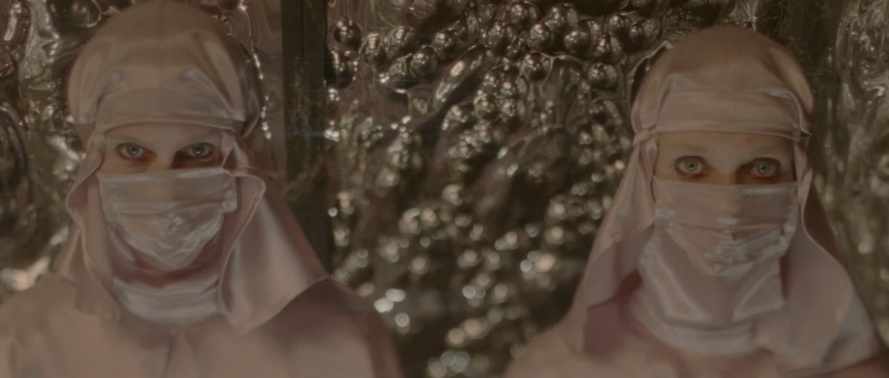 She Is Conann (2023) movie still: medium — Two identical figures wearing pink satin headgear stand before a textured, reflective silver wall. The…