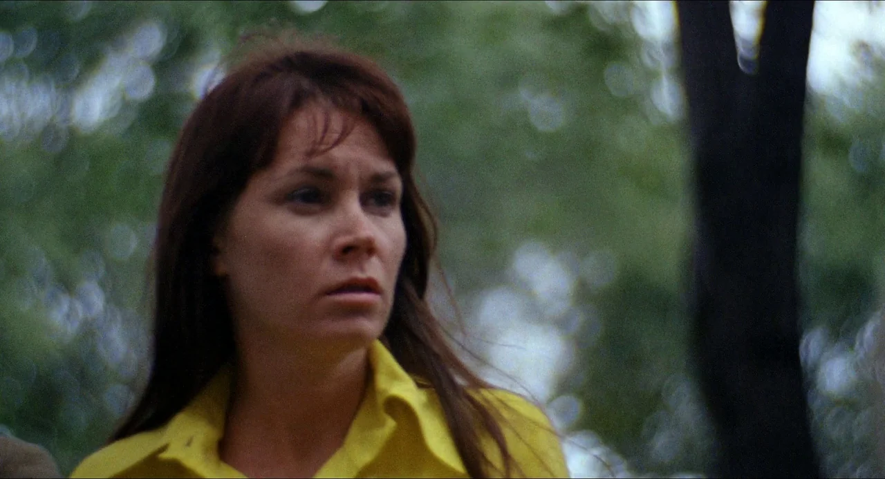 Medium Cool (1969) movie still: closeup — A woman stands in a lush outdoor area with out-of-focus trees and foliage behind. She is wearing a yellow…