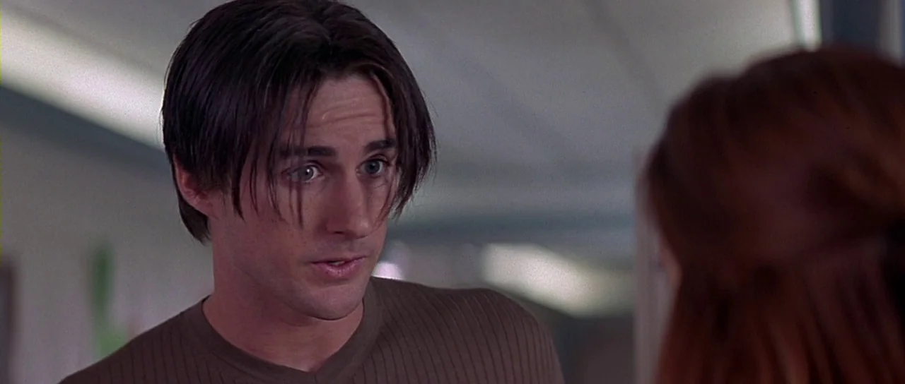 Scream 2 (1997) movie still: closeup — An interior hallway with light-colored walls and fluorescent ceiling lights. A man with brown hair talks to…