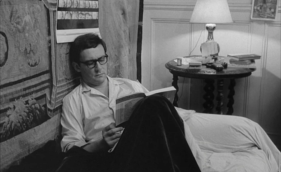 Le Feu Follet (1963) movie still: medium — The setting is a bedroom with a tapestry on the wall behind the subject. A man sits on a bed reading an…