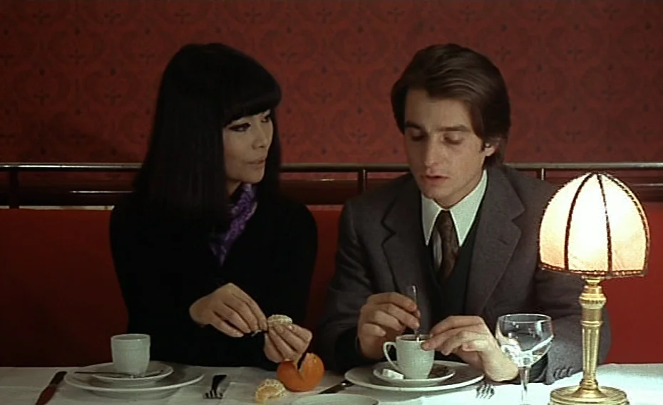 Bed & Board (1970) movie still: medium — A restaurant booth with red patterned wallpaper and red bench seating. A man and woman sit opposite each…