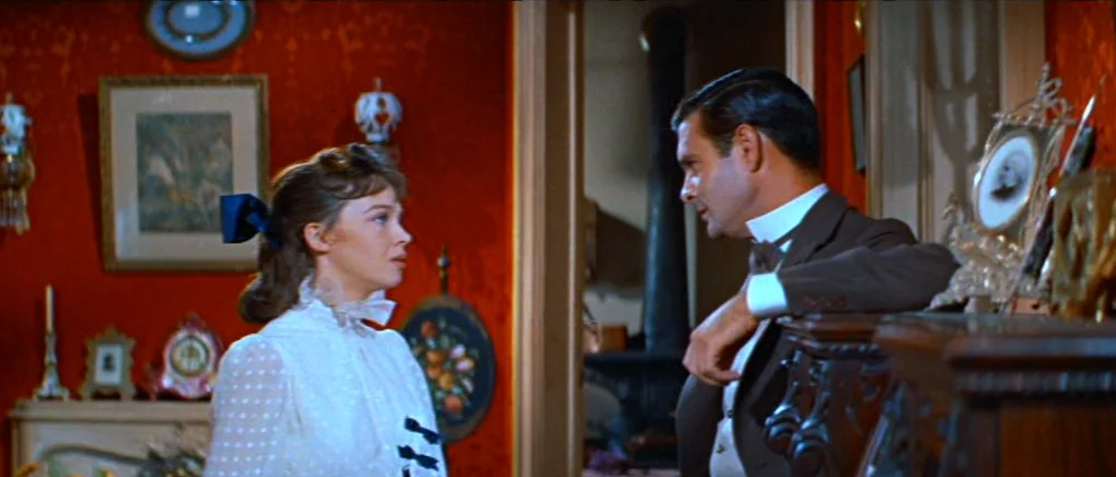 Gigi (1958) movie still: medium — A parlor with vibrant red patterned wallpaper adorned with framed artwork and decorative plates. A woman in a…
