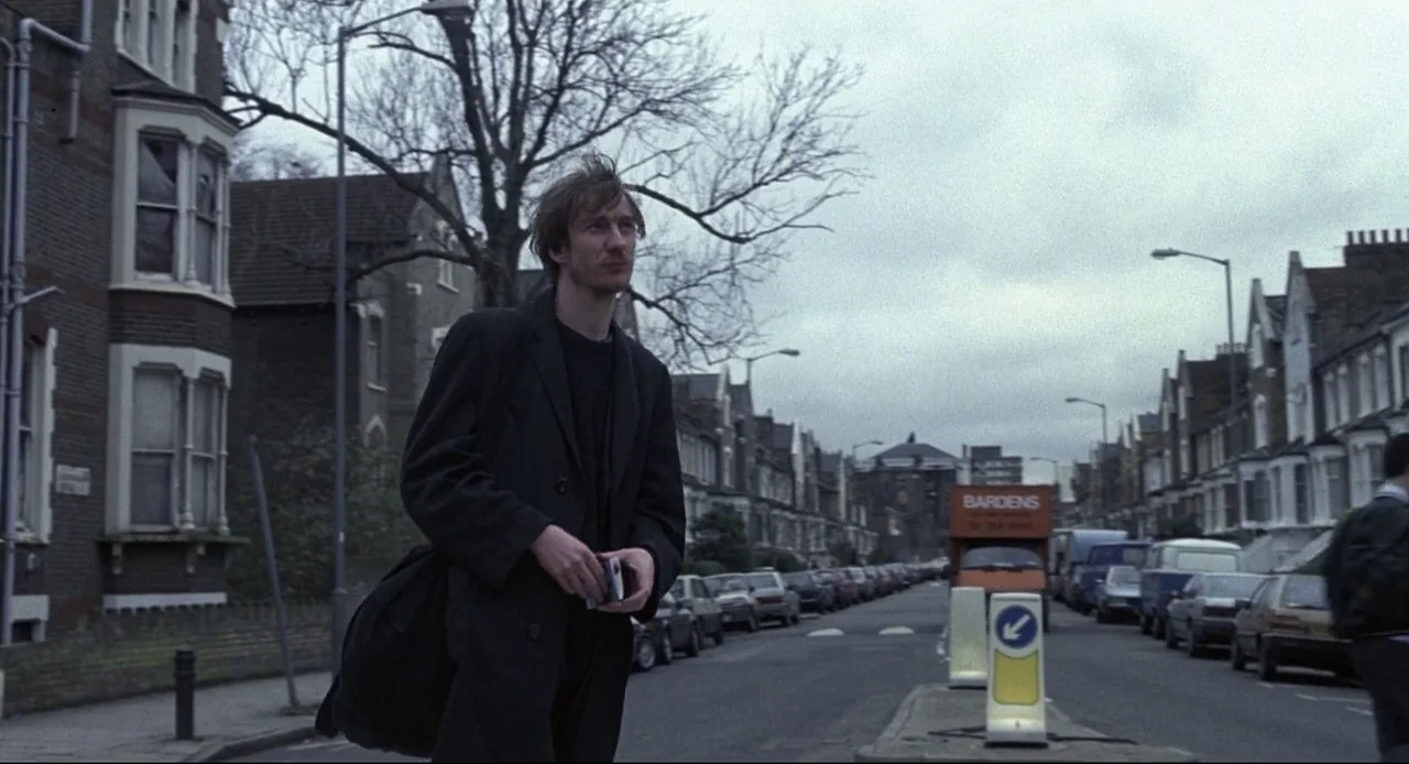 Naked (1993) movie still: medium — A residential British city street is lined with multi-story Victorian terrace houses. A thin man in a black coat…