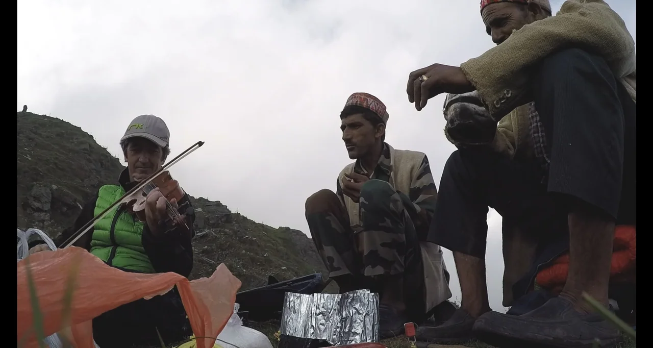 Blutch (2018) movie still: medium — A grassy mountain slope under a grey, overcast sky. Three men sit on the ground, with one playing a violin near…