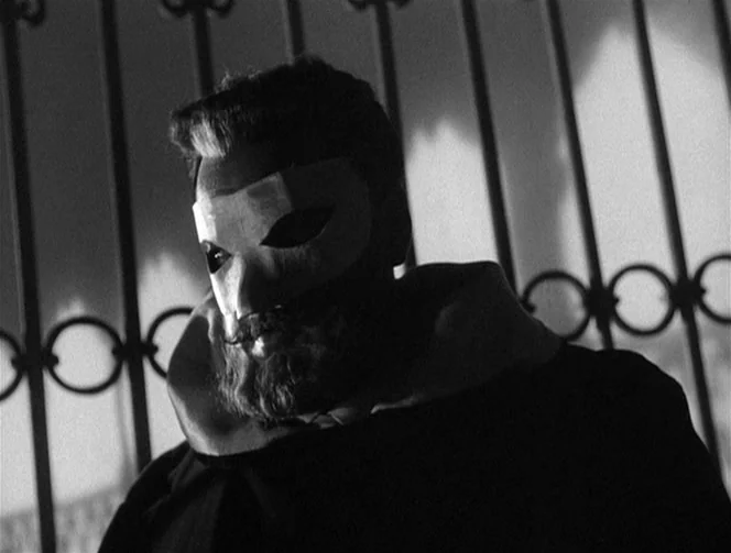Mr. Arkadin (1955) movie still: closeup — A man wearing a smooth mask and a dark hood stands before a gate. The metal bars are illuminated against a…