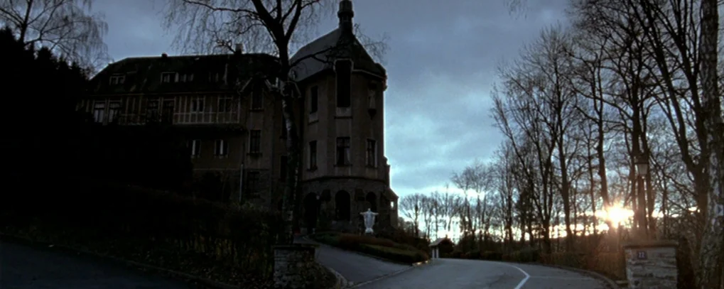 Calvaire (2004) movie still: establishing — A large stone manor stands at the end of a paved driveway at twilight. Bare trees surround the property…