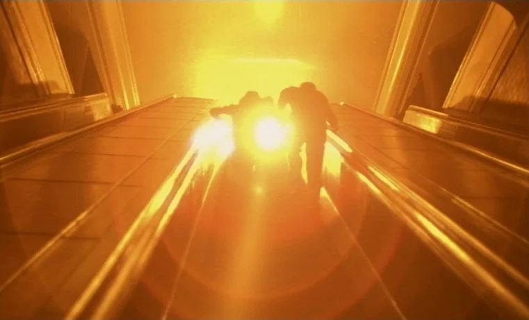 Golem (1980) movie still: wide — An interior escalator in a building glows with intense, blinding yellow artificial light. Two male figures walk…