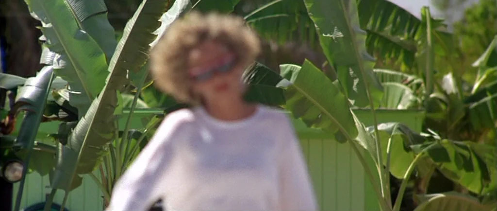 Manhunter (1986) movie still: medium — A woman stands in front of a lime green building wall surrounded by banana leaves. The foreground subject is…