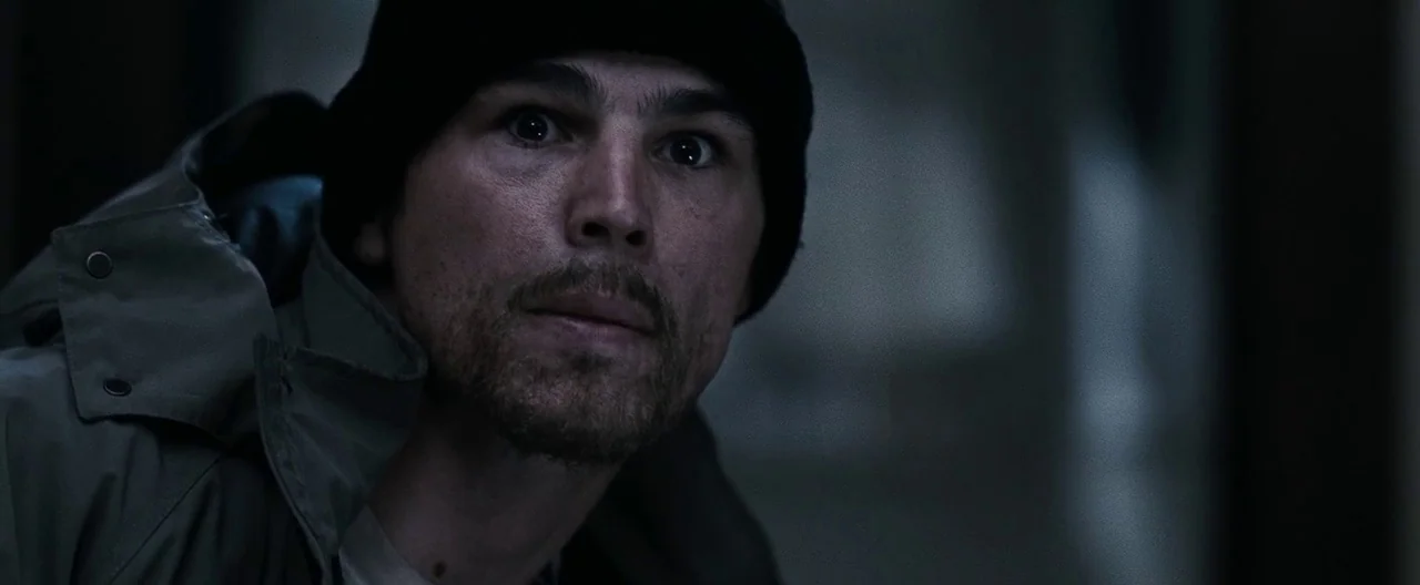 30 Days of Night (2007) movie still: closeup — A man stands in a dimly lit, nondescript interior space with dark walls. He stares forward with an…