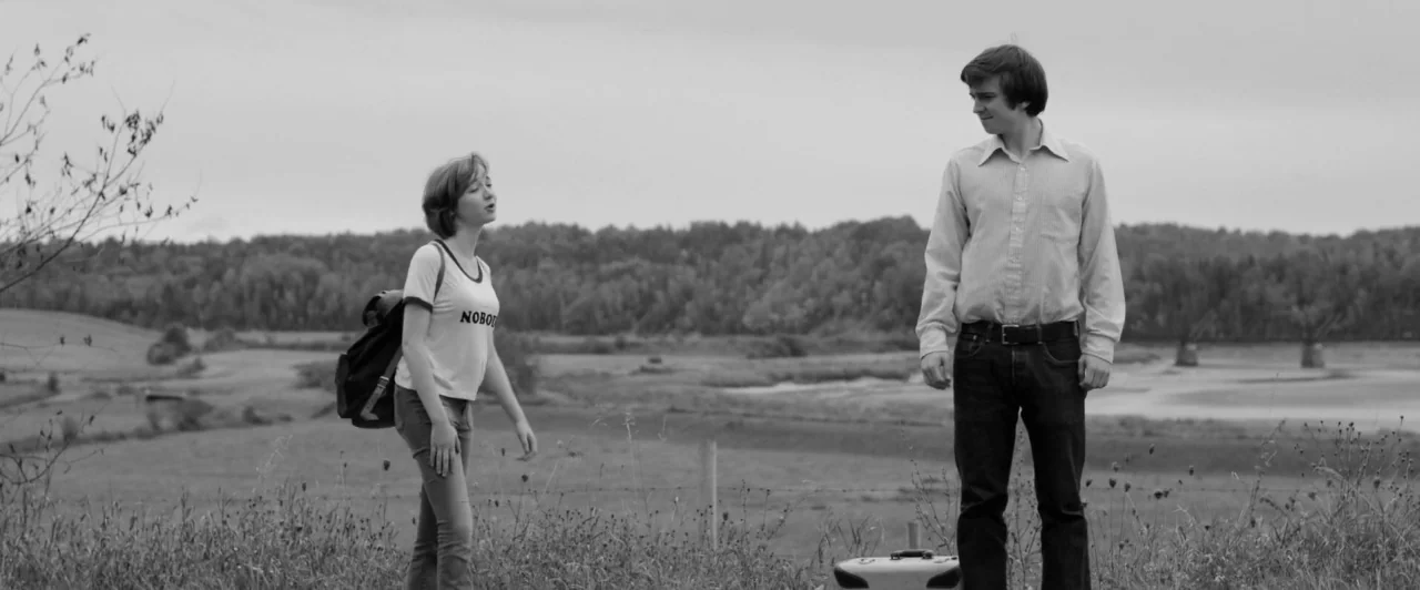 Weirdos (2016) movie still: fullbody — An open rural field under an overcast sky features sparse vegetation and distant trees. A teenage girl…