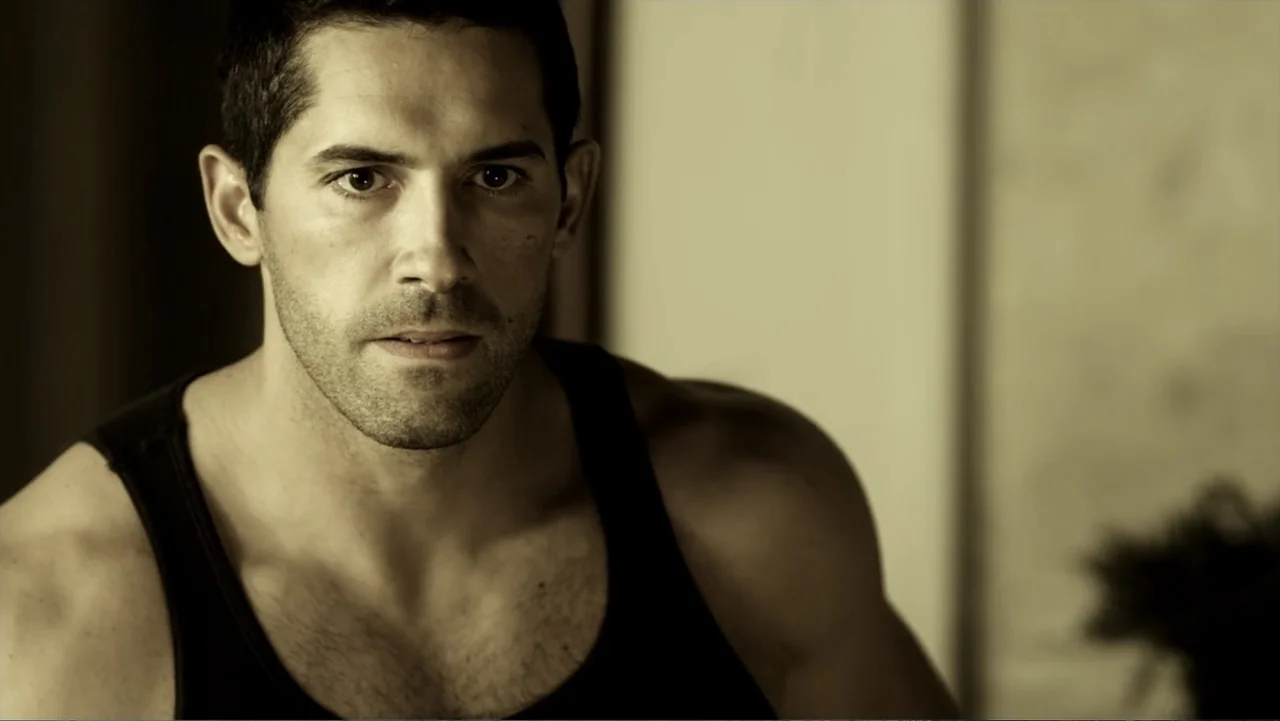 Assassination Games (2011) movie still: closeup — A man stands in an interior room with a plain wall behind him. The man looks directly into the…