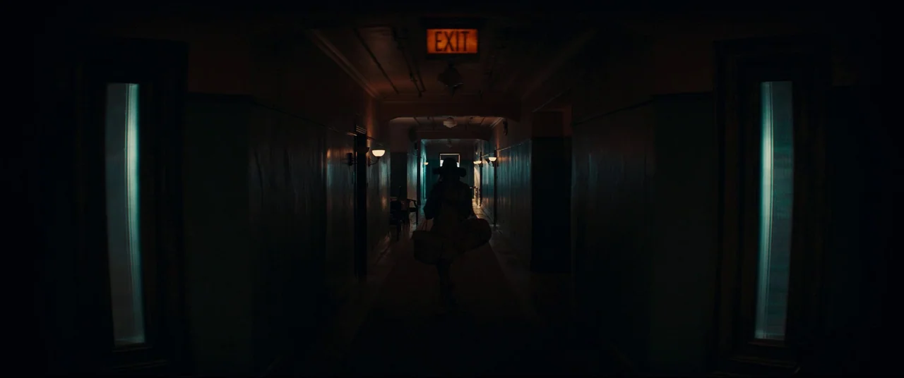 Evil Dead Rise (2023) movie still: wide — A long, shadowed hotel corridor stretches into the distance with blue-tinted walls. A single figure walks…