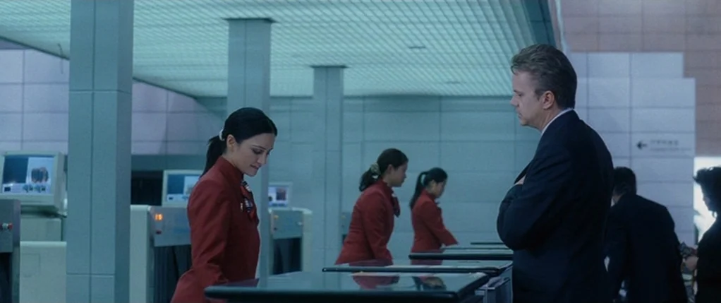 Code 46 movie still: medium — An airport security area with tiled walls and blue-toned lighting panels overhead. A man in a suit stands at a…