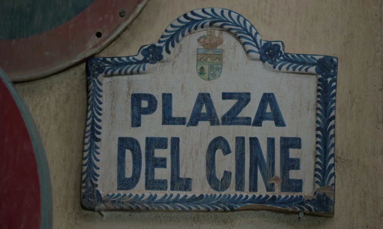 Close Your Eyes (2023) movie still: closeup — A ceramic street sign reading PLAZA DEL CINE is mounted on a textured wall. The sign features blue…