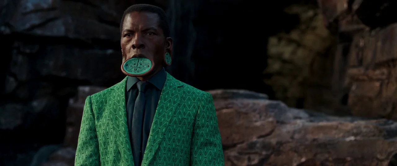 Black Panther (2018) movie still: medium — A man stands in front of dark, textured volcanic rocks in an outdoor setting. The subject wears a…