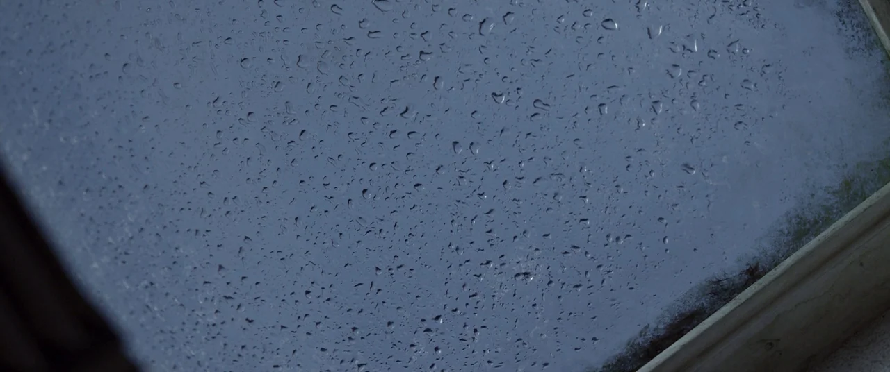 Room (2015) movie still: closeup — A close view of a glass windowpane covered in a layer of raindrops. The exterior beyond the glass is obscured and…