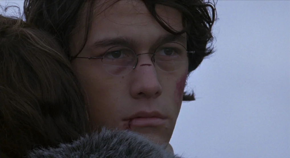 Brick (2005) movie still: closeup — A featureless outdoor space under a pale overcast sky during the day. A young man with visible facial injuries…