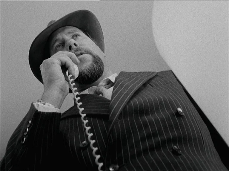The American Soldier (1970) movie still: medium — A man stands in an enclosed area talking on a vintage coiled telephone. He wears a pinstriped suit…