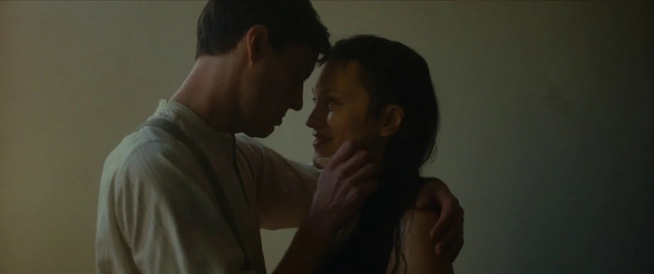 Earwig (2022) movie still: medium — An interior space with dim lighting and plain walls serves as the backdrop. A man in a shirt touches a woman's…