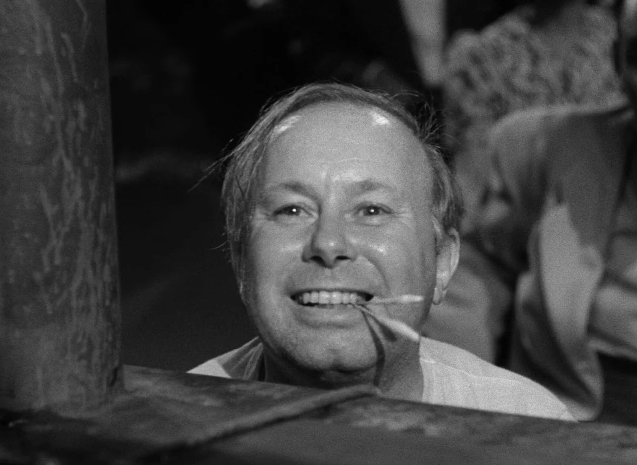 The Set-Up (1949) movie still: closeup — A dimly lit indoor space with a dark foreground structure framing the subject. The man smiles directly at…