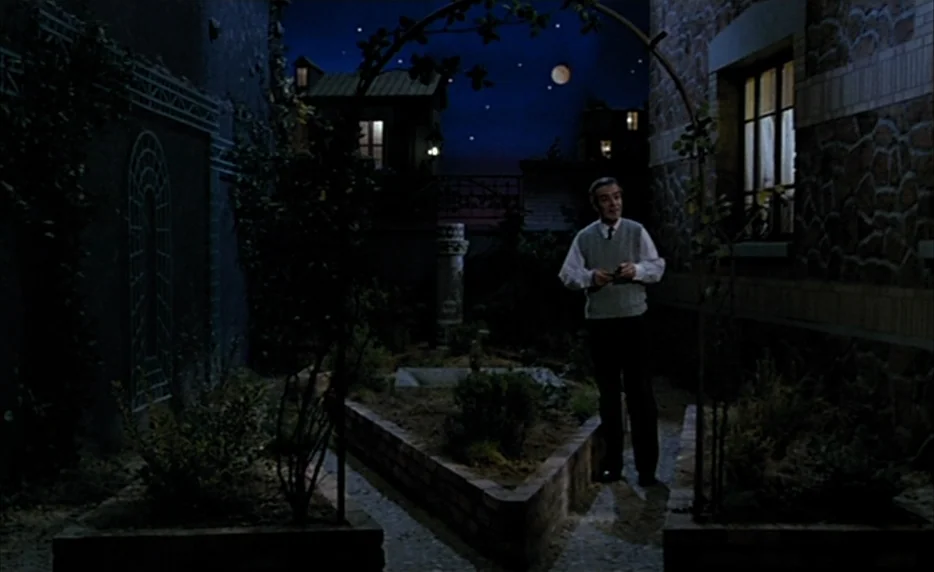 Mélo (1986) movie still: fullbody — A night time courtyard features stone walls, raised planting beds, and a metal trellis arch. A middle aged man…