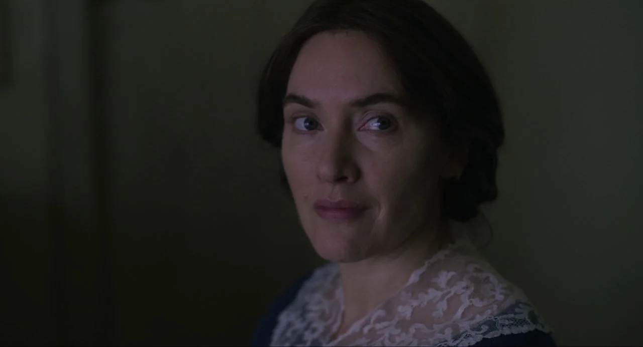 Ammonite (2020) movie still: closeup — A dimly lit room provides a dark, muted background for a person. A woman with dark hair and a lace-trimmed…