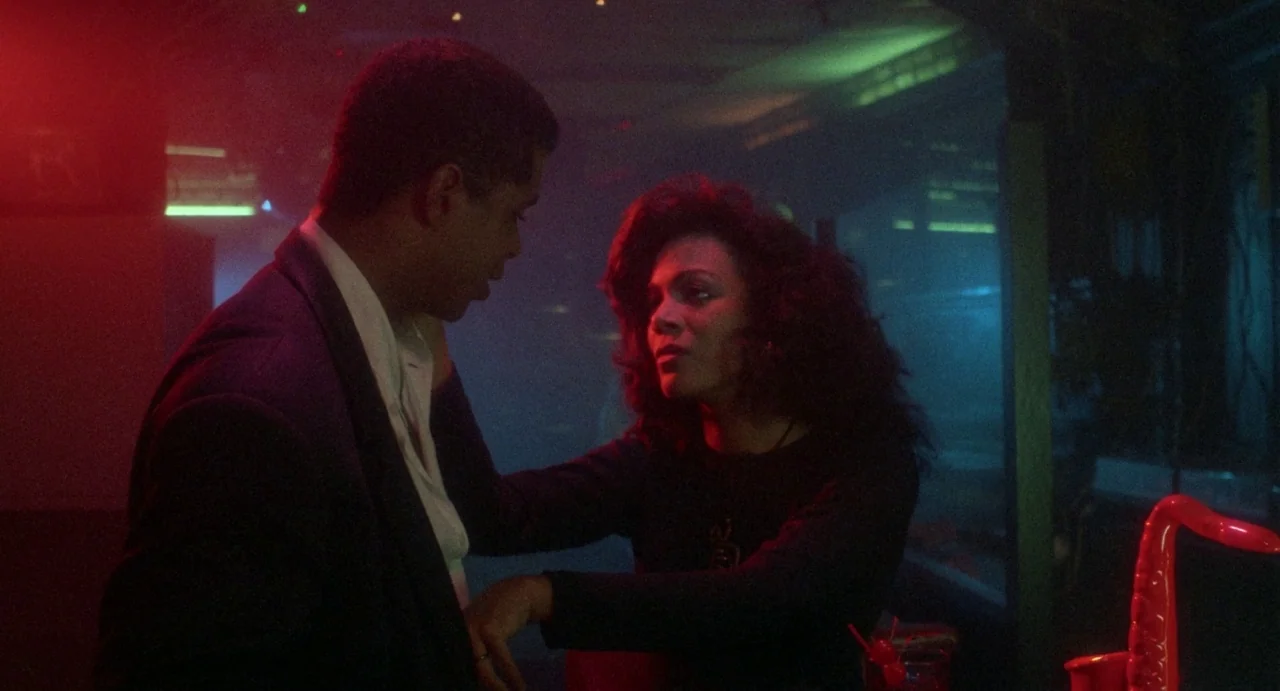 Def By Temptation (1990) movie still: medium — A dimly lit nightclub with hazy air filled with neon red and green light. A woman in a black shirt…