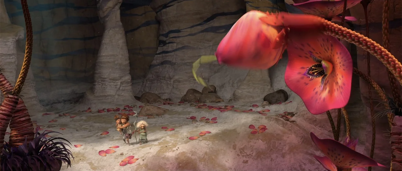 The Croods (2013) movie still: wide — A prehistoric cave environment features sandy ground scattered with fallen pink flower petals. Two adults and…