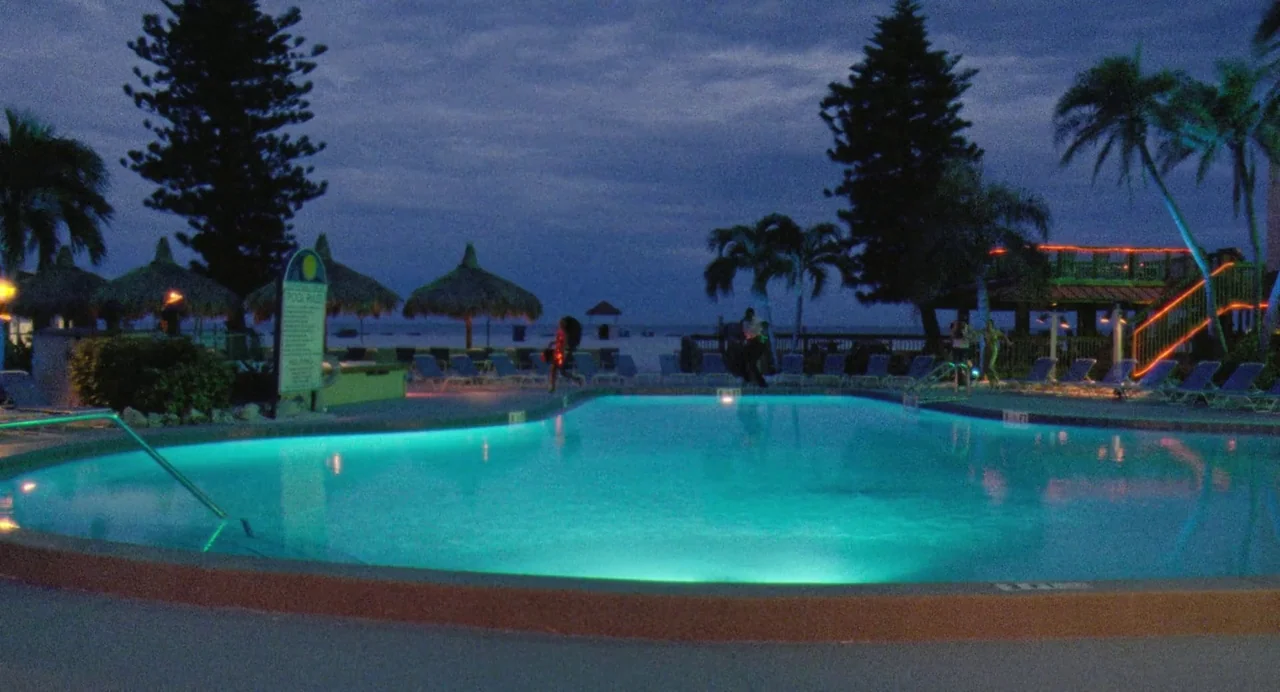 Zola (2020) movie still: wide — A resort swimming pool glows with turquoise underwater lights at night under a dark sky. Several palm trees and…