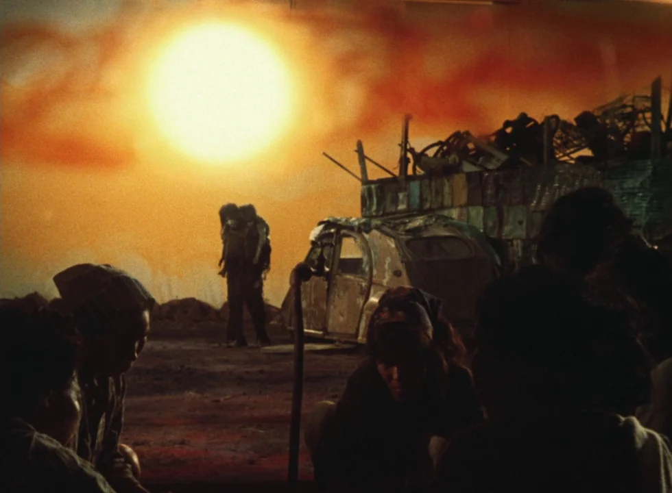 Dodes’ka-den (1970) movie still: wide — A desolate, debris-strewn wasteland stretches out under a massive, hazy orange sun. A person carries another…