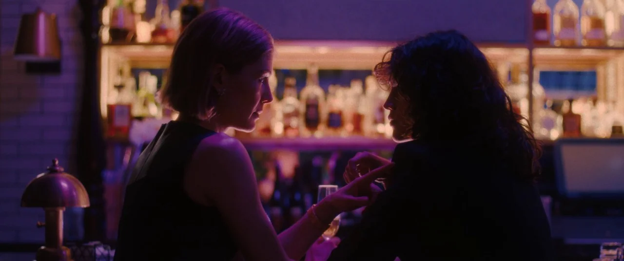 I Care A Lot (2020) movie still: medium — A dimly lit bar features shelves of glass bottles glowing under vibrant neon light. Two women sit at the…