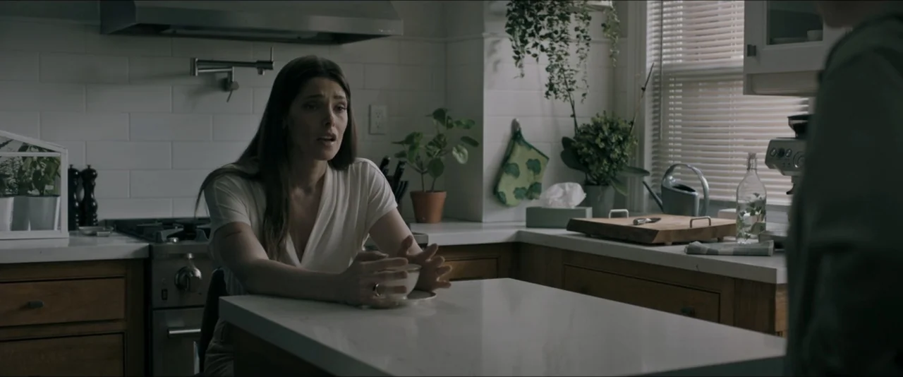 It Feeds (2025) movie still: medium — A residential kitchen features white subway tile, wood cabinets, and a white stone island. A woman sits at the…