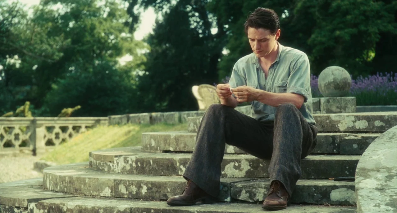 Atonement (2007) movie still: fullbody — A man sits on weathered stone steps in a verdant estate park area. He focuses intently on rolling a…