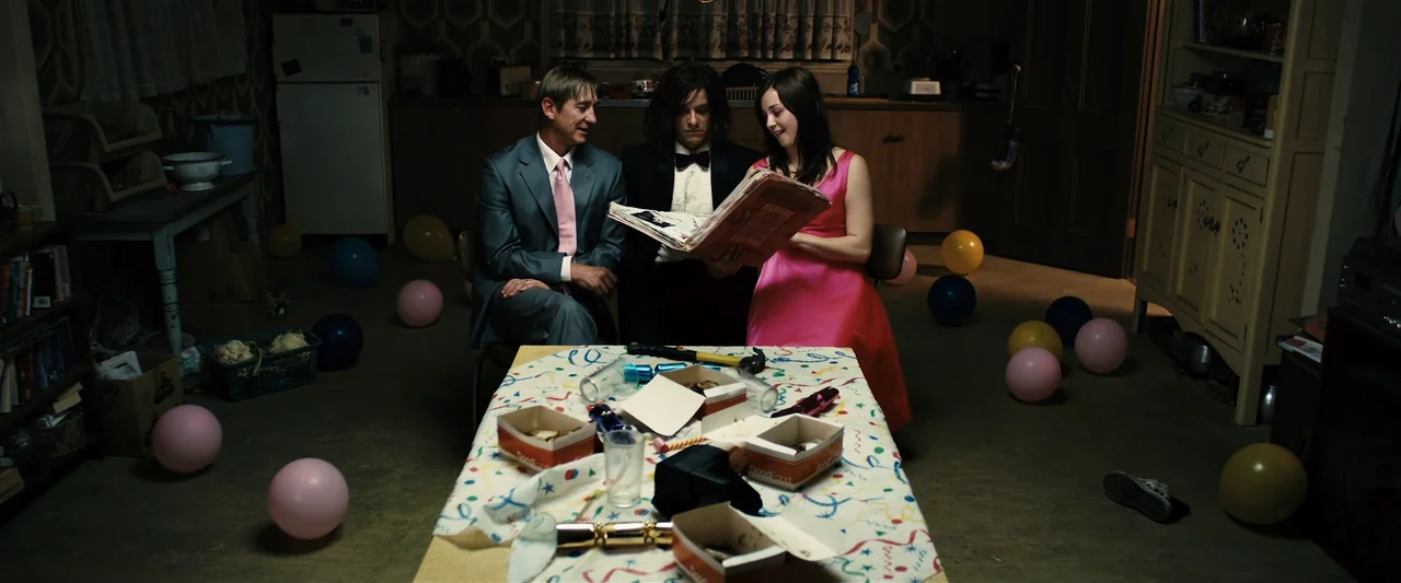 The Loved Ones (2009) movie still: wide — A dimly lit residential interior features patterned wallpaper and a confetti-covered dining table. Three…