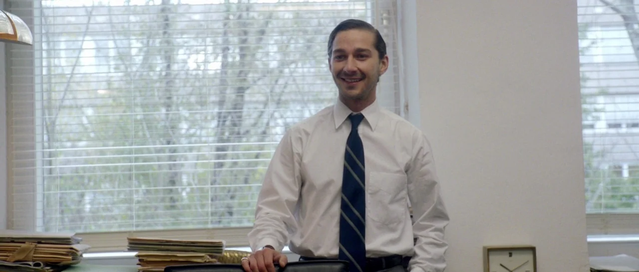 Nymphomaniac Vol 1 (2013) movie still: medium — A bright workspace features large windows with white horizontal blinds showing trees outside. A man…