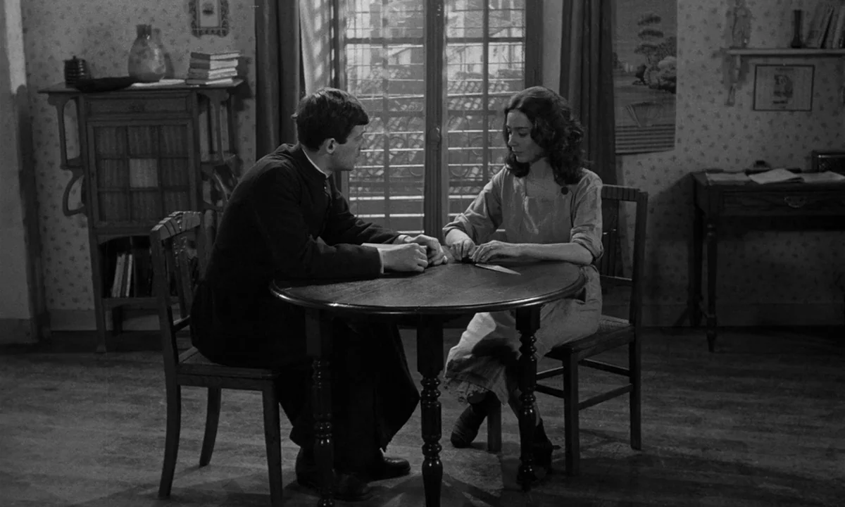 Léon Morin, Priest (1961) movie still: medium — A spare, wood-paneled room features floral wallpaper and a large central window. A young priest in a…