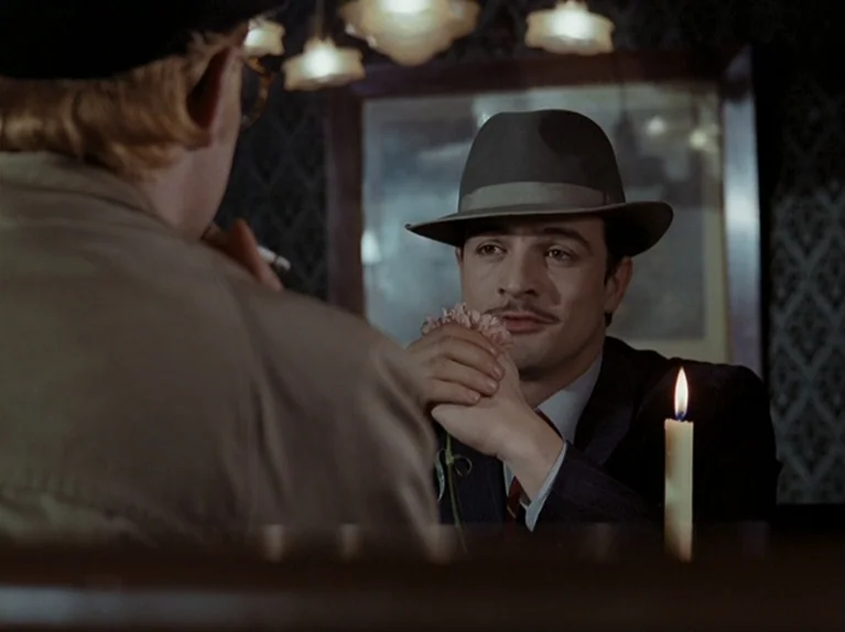 The Third Generation (1979) movie still: medium — A man in a fedora sits across from another person at a dimly lit table. He holds a small flower…