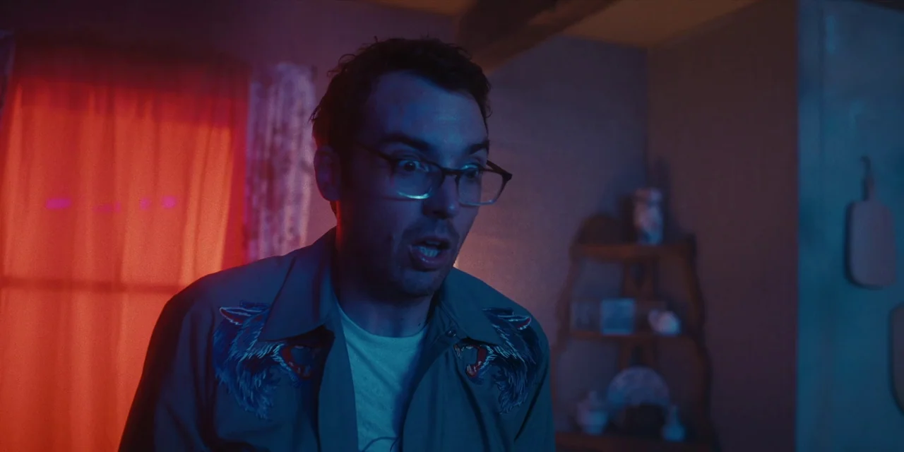 Destroy All Neighbours (2024) movie still: medium — A dark room illuminated by saturated red and blue light sources. A man wearing glasses looks…