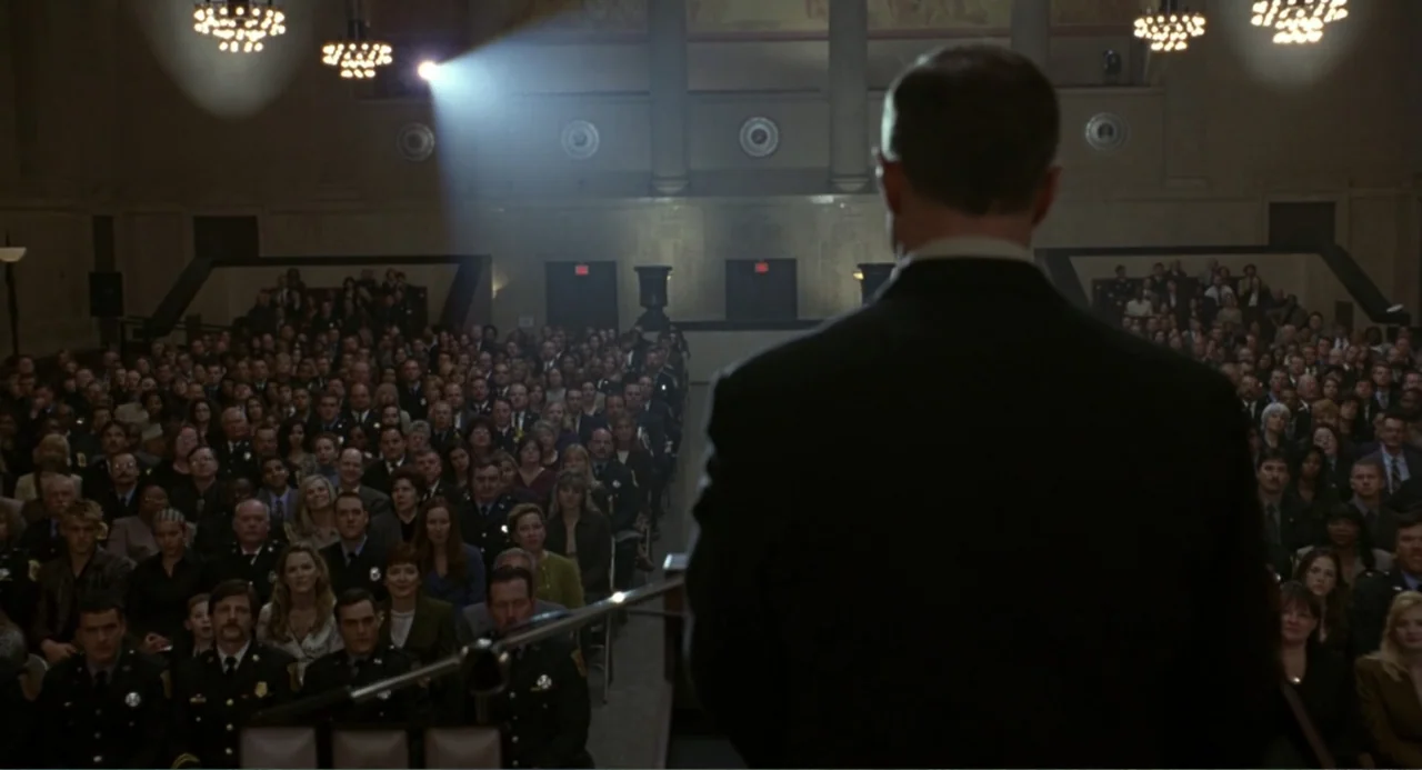 Ladder 49 (2004) movie still: medium — A dark auditorium holds a large audience seated in rows facing a lit podium. A man in a dark suit stands at…