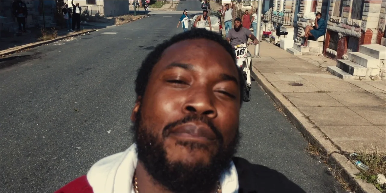 Charm City Kings (2020) movie still: closeup — An urban residential street lined with row houses and parked vehicles during the day. A man films…