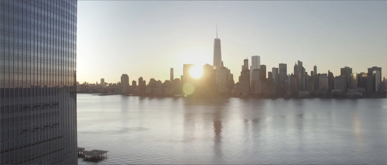 Square One (2019) movie still: establishing — The New York City skyline is visible across the Hudson River at sunrise. A modern glass skyscraper…