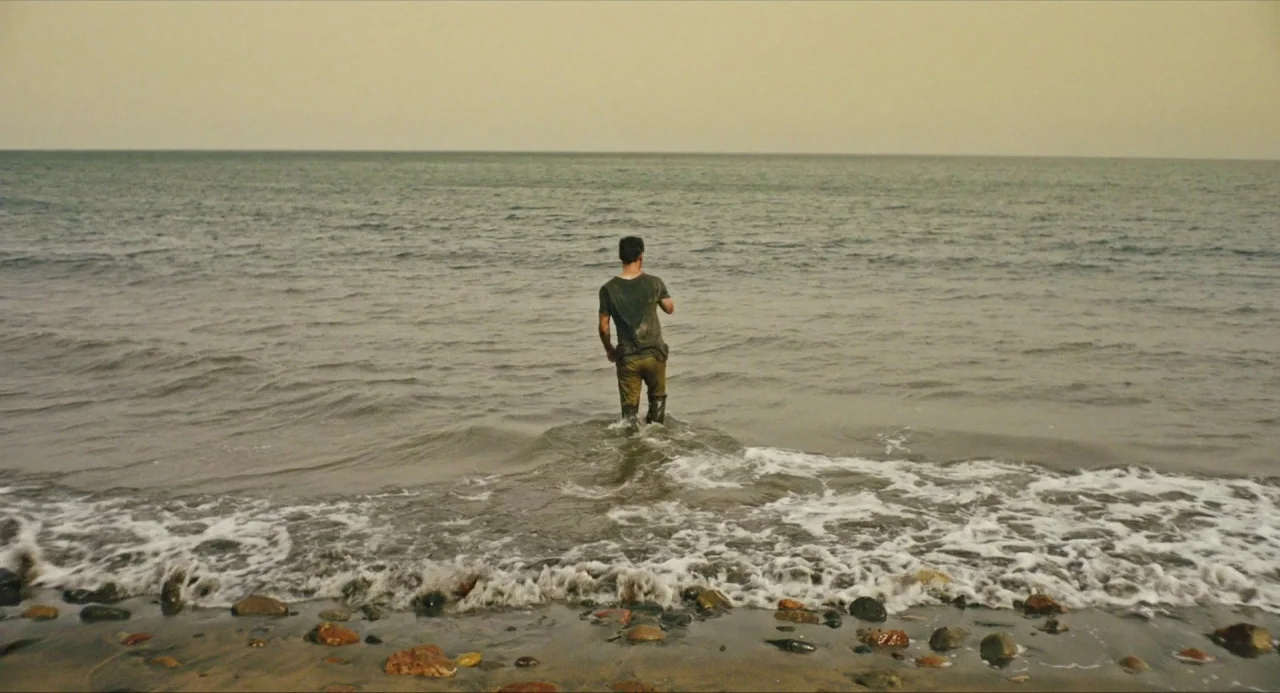 Submergence movie still: wide — A pebble-strewn beach meets a grey-green ocean under a hazy yellow-tinted sky. The man stands knee-deep in the…